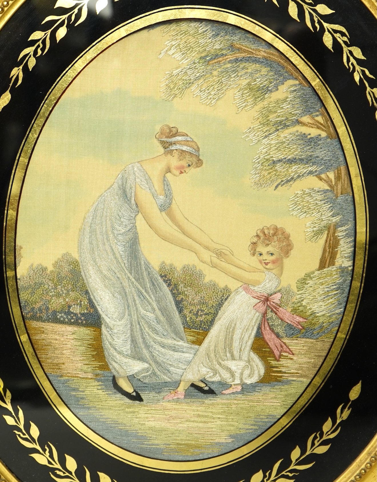 A pair of early 20th century oval silk embroideries, one of a mother and child dancing in a garden, the other of a lady in a large brimmed hat, watering flowers in by a classical stone urn, watched by her dog. Framed in